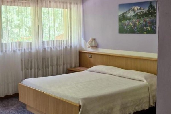 Jednoduše zařízená ložnice s manželskou postelí, obrázkem hory na zdi a závěsy u oken.