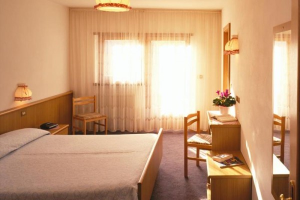 Světlý hotelový pokoj s postelí, nočními stolky, židlemi a toaletním stolkem vedle okna se záclonami.