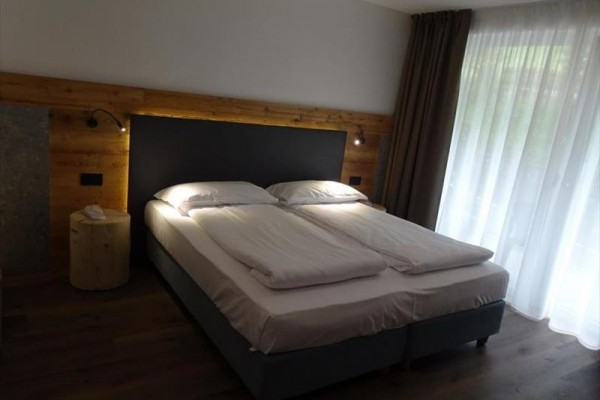 Manželská postel v moderní ložnici s dřevěnými prvky a velkým oknem.