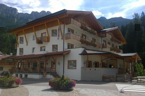 Třípodlažní horský hotel obklopený horami a lesem s květinovými truhlíky na balkonech.