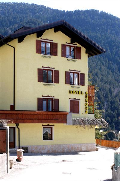 Žlutý hotel s hnědými okenicemi před zalesněným kopcem.  