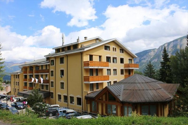 Budova hotelu v horském prostředí s parkovištěm a auty v popředí.