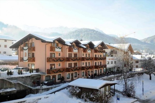 Zasněžený hotel s balkonky a horami v pozadí.