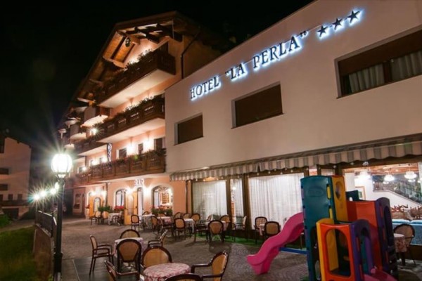 Noční pohled na osvětlený hotel s venkovním posezením a dětským hřištěm.