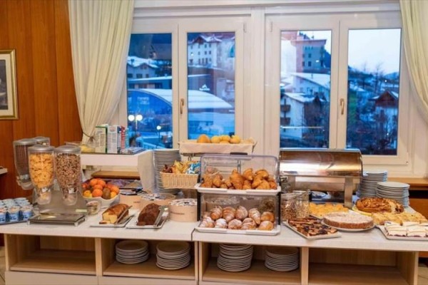 Bufetový stůl s pečivem, croissanty, cereáliemi a ovocem před okny s výhledem na zasněžené město.