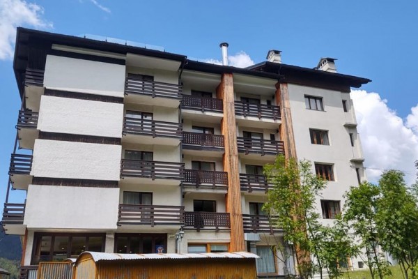 Vícepodlažní budova s balkony obklopená stromy pod modrou oblohou.