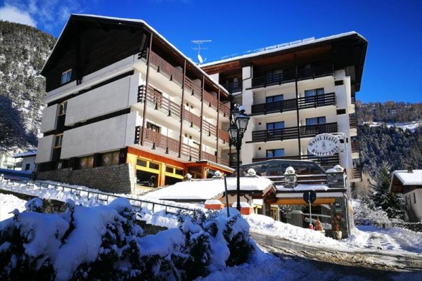 Zasněžený horský hotel s dřevěnými balkony za slunečného dne.