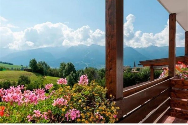 Dřevěný balkon s květinami a výhledem na hory s modrou oblohou a oblaky.