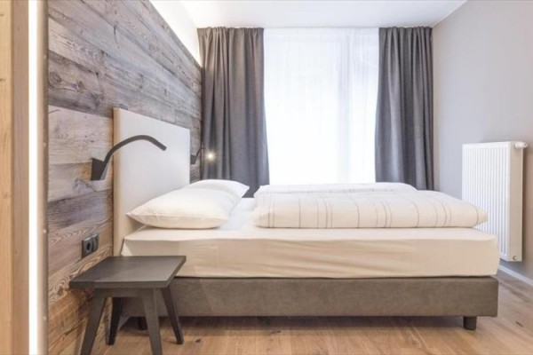 Moderní ložnice s dřevěnou stěnou, dvojlůžkem a tmavými závěsy.