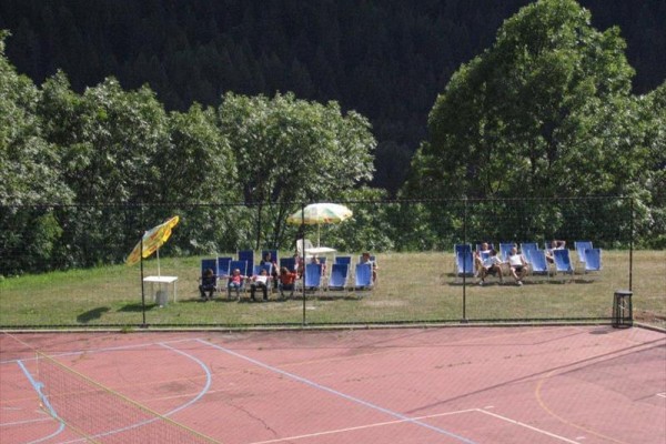 Lehátka se slunečníkem na trávníku vedle prázdného sportovního hřiště obklopeného stromy.