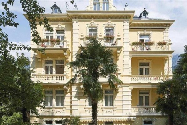 Žlutá secesní budova s balkony, obklopená palmami a stromy.  