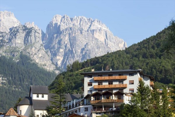 Hotel s dřevěnými balkony na úpatí zalesněné hory s skalnatými vrcholy.