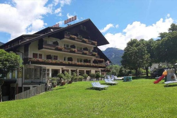 Hotel s balkony plnými květin, před nímž je rozlehlý travnatý prostor s lehátky a skluzavkou.