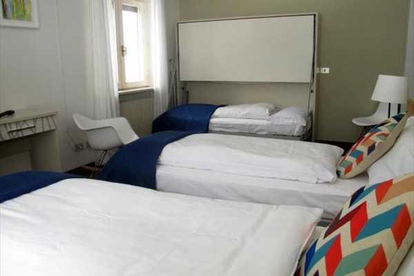 Tři postele v moderně zařízeném pokoji s bílými stěnami a barevnými polštáři.