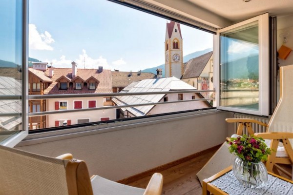 Výhled z balkonu s dřevěnými křesly na zvonici kostela a horské panorama.