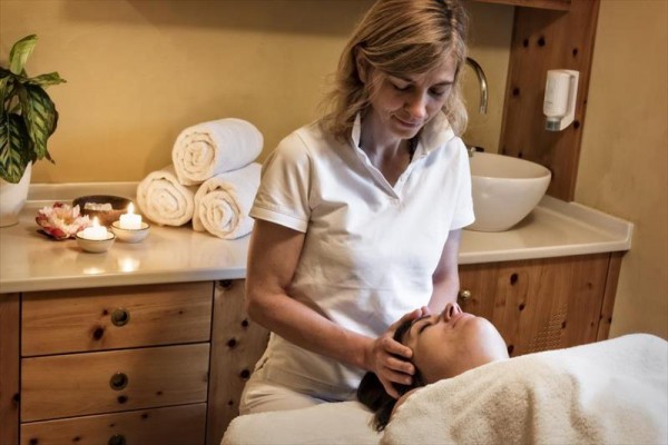 Žena v bílém provádí masáž hlavy v relaxační místnosti s tlumeným osvětlením a svíčkami.