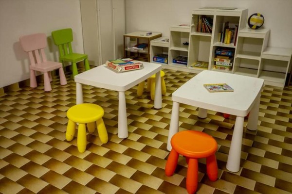 Dětská herna s barevnými židličkami a stolky na šachovnicové podlaze.