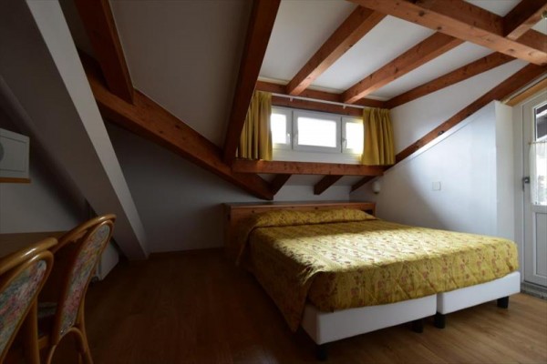 Podkrovní ložnice s dřevěnými trámy a oknem se žlutými závěsy.