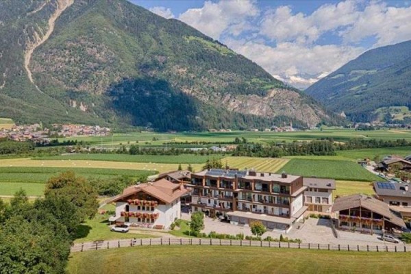 Hotel obklopený zelenými poli a horami na obzoru pod modrou oblohou s obláčky.  