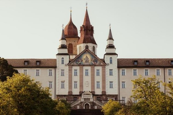 Historická budova s věžemi a freskou na průčelí obklopená stromy.