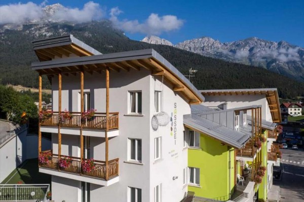 Moderní horský hotel se zelenou a bílou fasádou, s pozadím hor a oblohy s oblačností.