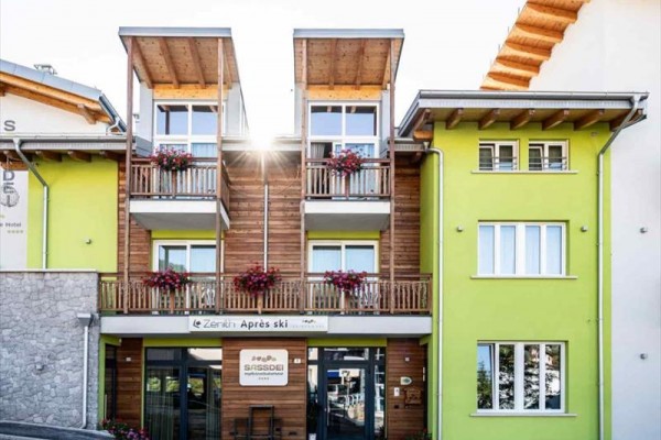 Budova s dřevěnými balkony a květinami na fasádě, vedle zeleně natřeného domu.  