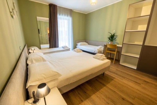 Pokoj s dvěma postelemi, psacím stolem, otevřenou policí a závěsy u okna.