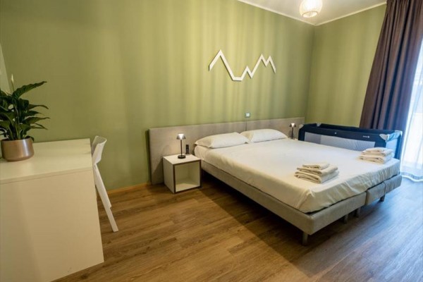 Moderní ložnice se zelenými stěnami, manželskou postelí, dětskou postýlkou a minimalistickým nábytkem.