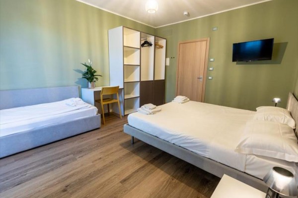 Moderní hotelový pokoj s manželskou postelí, jednolůžkem, skříní a televizí na stěně.
