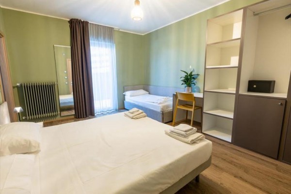 Světlý pokoj s manželskou postelí, přistýlkou, policemi, stolem, židlí a rostlinou.
