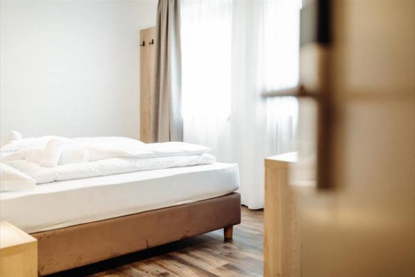 Světlá ložnice s postelí, bílým povlečením a dřevěnou podlahou.