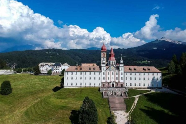 Historická budova s věžemi na pozadí hor a modré oblohy s mraky.