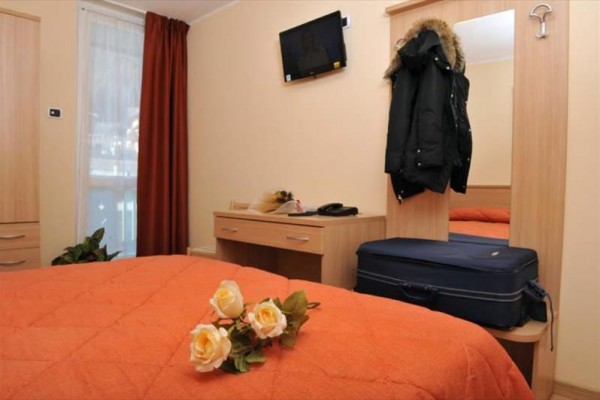 Hotelový pokoj s oranžovým přehozem na posteli, růžemi a zavazadlem u zrcadla.