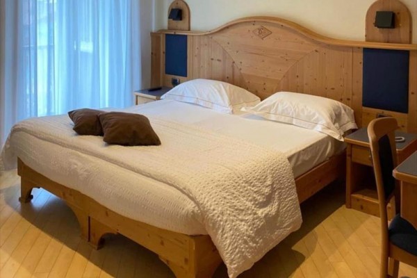 Dřevěná postel v ložnici s bílým povlečením a dvěma polštáři, vedle noční stolek a okno se závěsem.