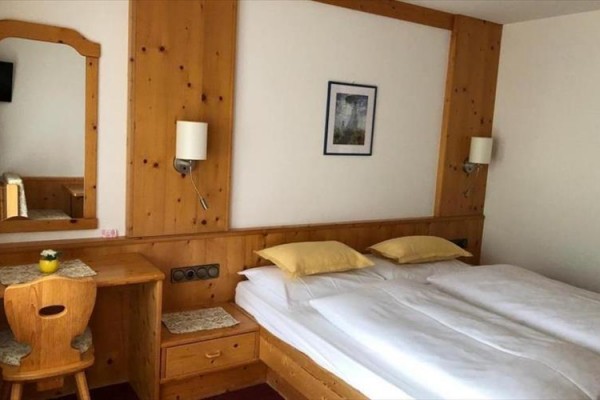 Hotelový pokoj s dřevěným nábytkem, manželskou postelí a psacím stolem se zrcadlem.  