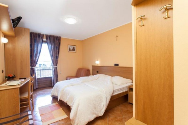 Pokoje s dvoulůžkem, psacím stolem a balkonem, na zdi visí kříž.